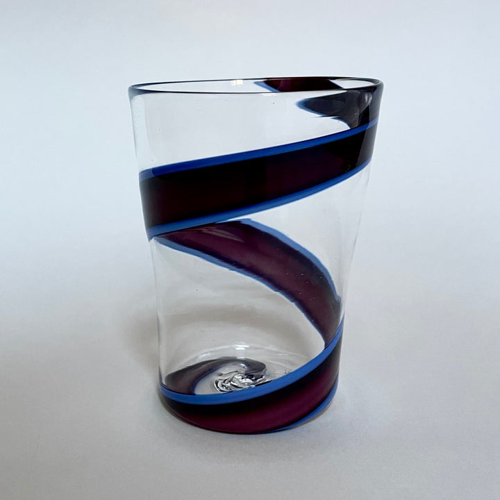 A glamorous addition to everyday table, this mouth blown Murano glass will elevate any table setting with its dynamic and colorful look.  An enveloping, black swirl accents the sleek, tumbler shaped silhouette, effortlessly dancing on the crystal clear surface enriched with 24 K gold leaf added during the blowing process.  Part of the Vortex Collection in different colors, this glass will complement any modern or classic decor with elegance.